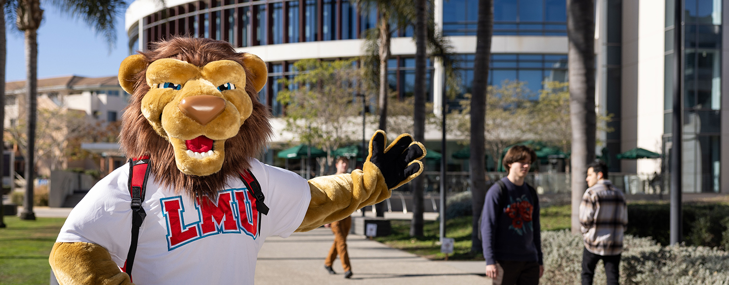 The Iggy Lion mascot, from the waist up, wearing an LMU t-shirt, standing at a distance in front of Hannon library on Palm Walk.